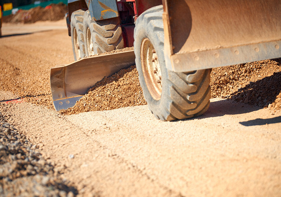 AdobeStock_257438510 Road Grader Spreading Gravel On road Construction Site - Closeup view